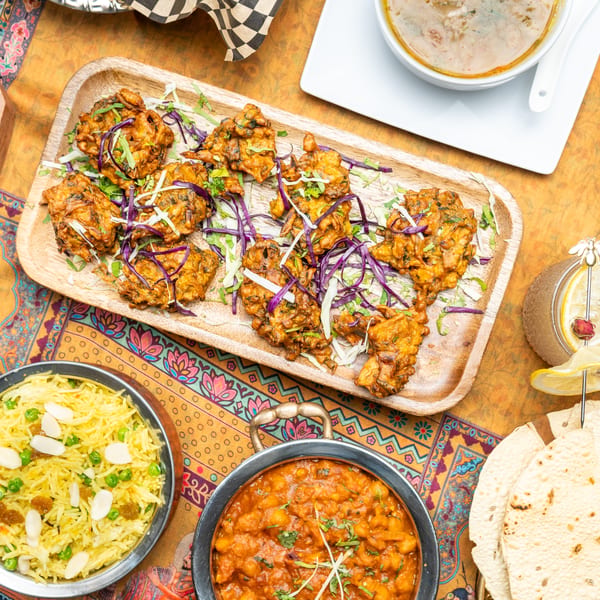Overhead view of Indian dishes including pakoras, rice, curry, soup, and papadum on a colorful tablecloth.