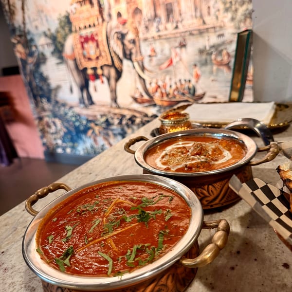 Two copper bowls of Indian curry, one red and one orange, on a marble counter with a mural.