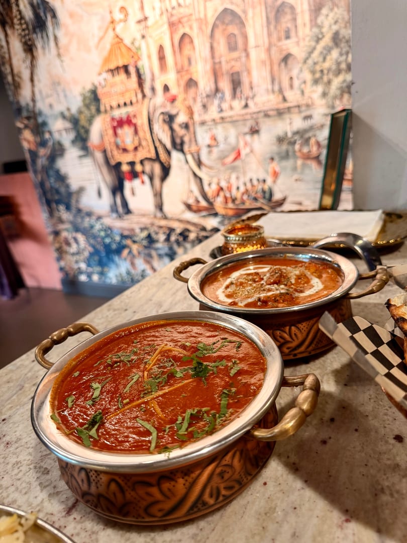 Two copper bowls of Indian curry, one red and one orange, on a marble counter with a mural.