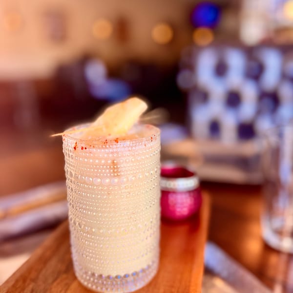 A creamy cocktail in a textured glass, garnished with a fruit slice and red spice, on a wooden tray.