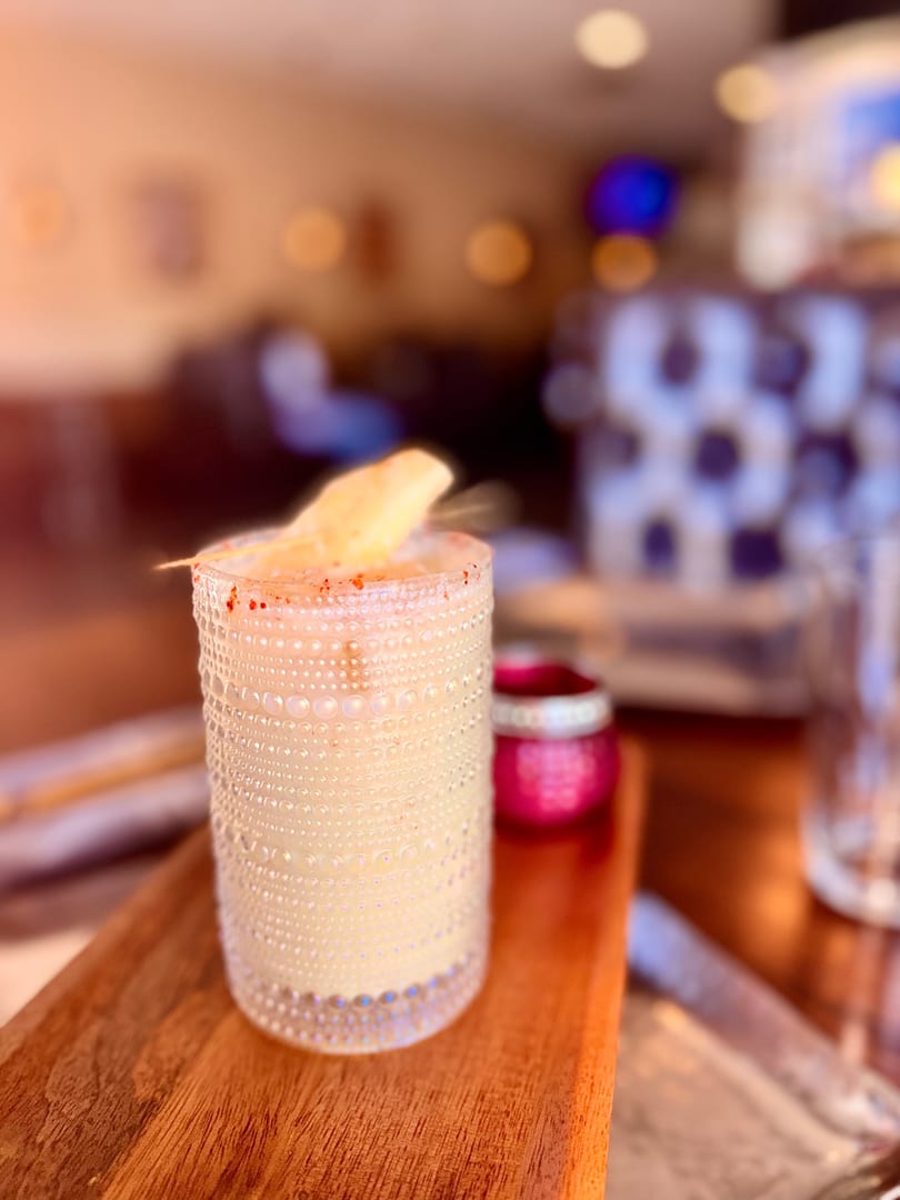 A creamy cocktail in a textured glass, garnished with a fruit slice and red spice, on a wooden tray.