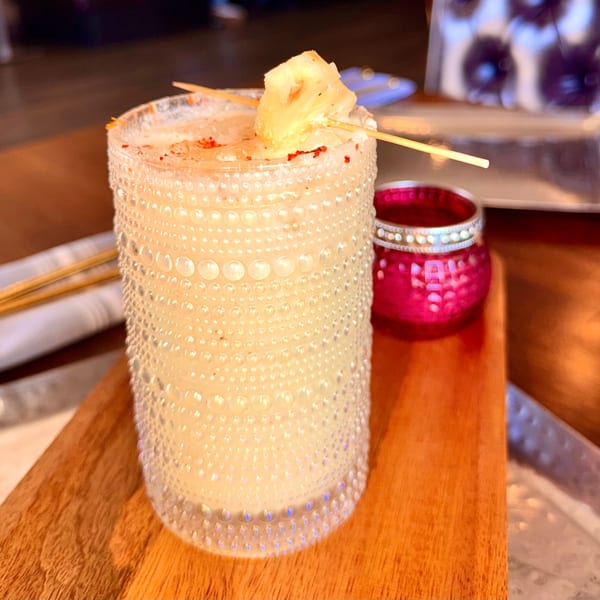 A textured glass of light-colored cocktail, garnished with pineapple and red spice, on a wooden board.