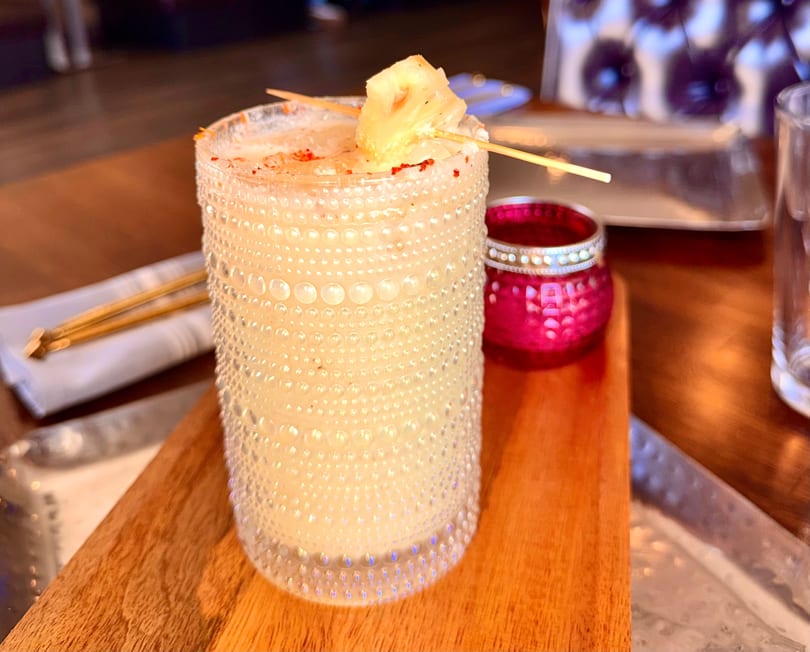 A textured glass of light-colored cocktail, garnished with pineapple and red spice, on a wooden board.