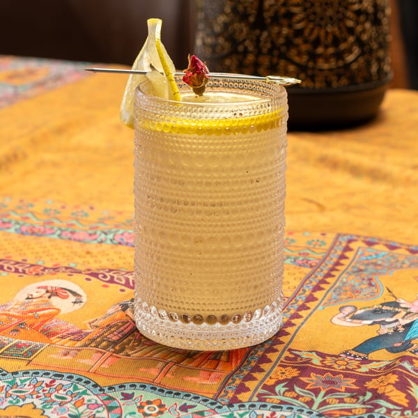 A cocktail in a textured glass, garnished with lemon and a dried rosebud, on a patterned yellow tablecloth.