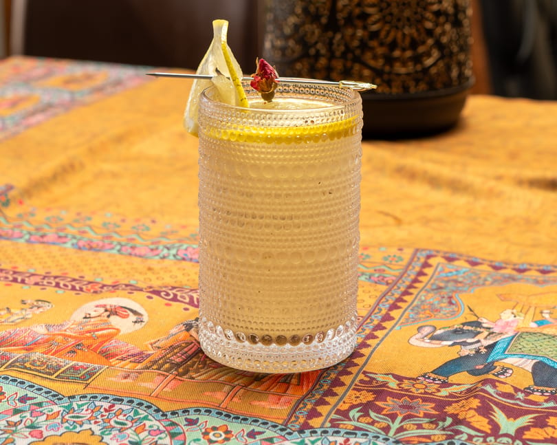 A cocktail in a textured glass, garnished with lemon and a dried rosebud, on a patterned yellow tablecloth.