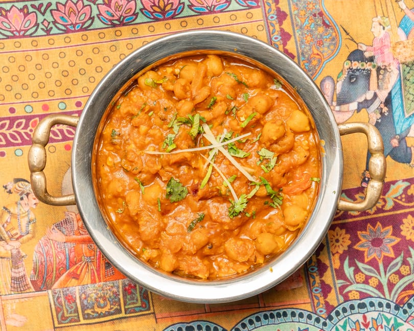 A metal bowl of chana masala, garnished with cilantro and ginger, on a colorful patterned tablecloth.