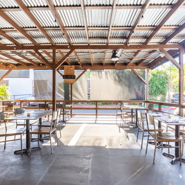 Outdoor patio with wooden beams, corrugated metal roof, tables, chairs, and sun shining through.