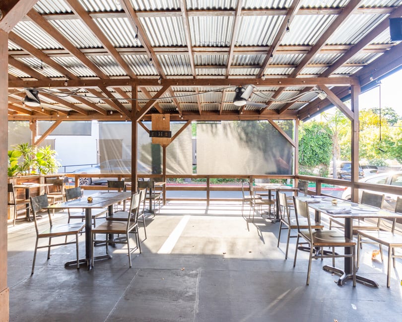 Outdoor patio with wooden beams, corrugated metal roof, tables, chairs, and sun shining through.