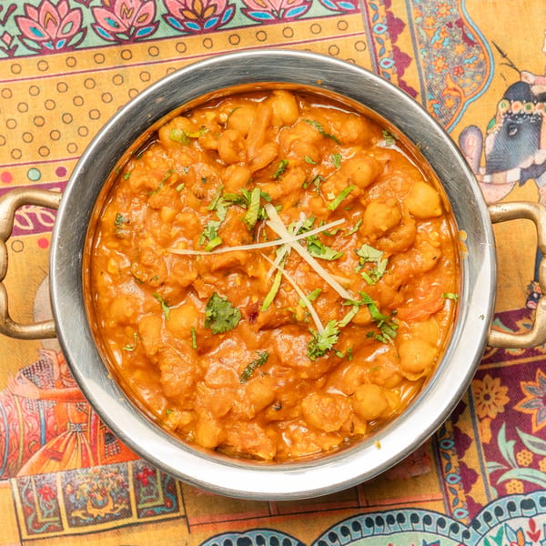 A metal bowl of chana masala, garnished with cilantro and ginger, on a colorful patterned tablecloth.