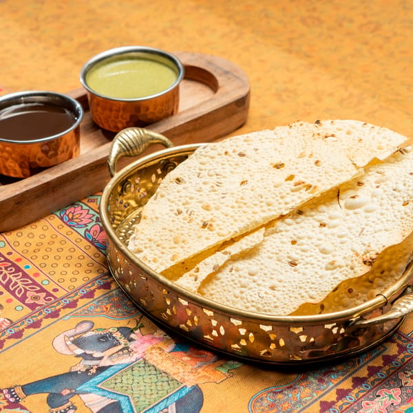 Crispy papadum in a brass bowl with two dipping sauces on a colorful patterned tablecloth.