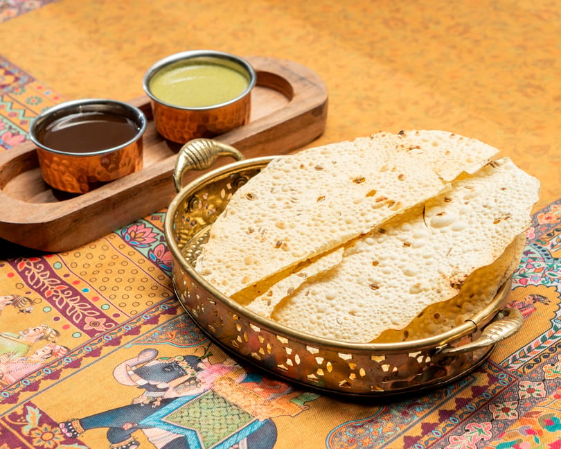 Crispy papadum in a brass bowl with two dipping sauces on a colorful patterned tablecloth.