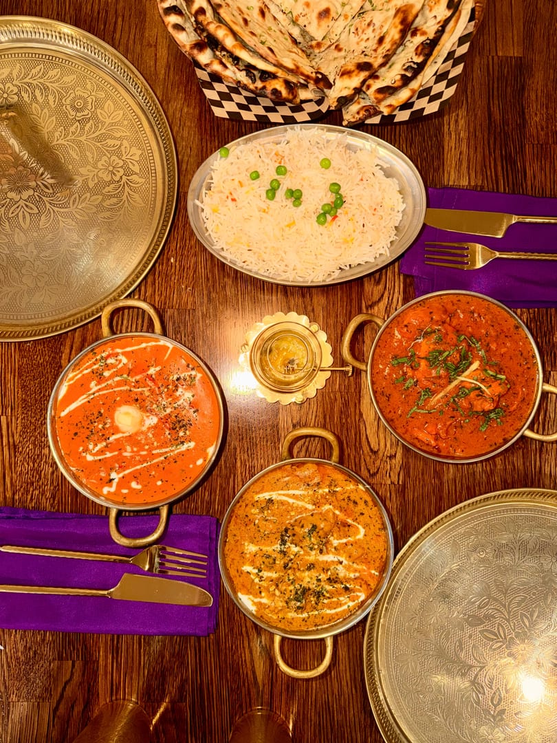 Overhead view of three Indian curries, rice, and naan bread on a wooden table.