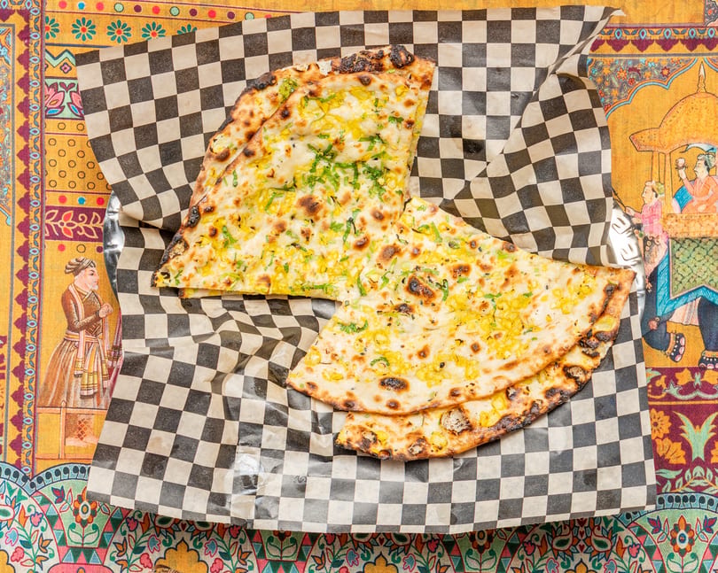 Two pieces of golden-brown naan bread with corn and herbs on checkered paper, on a colorful patterned tablecloth.