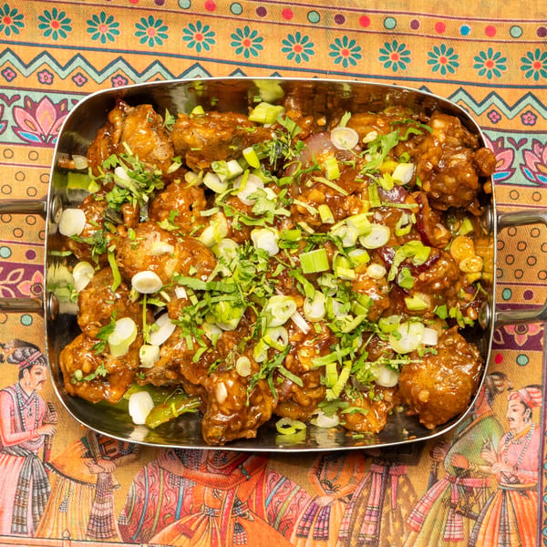Chili chicken in a metal serving dish, garnished with green onions and cilantro, on a colorful patterned tablecloth.