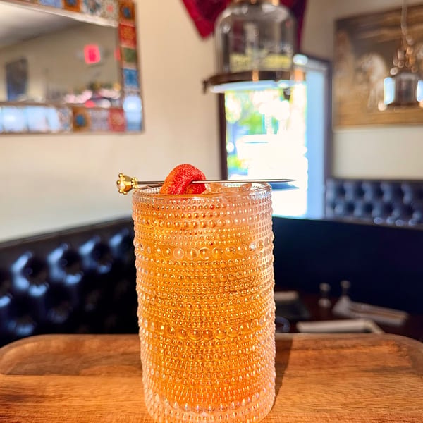 A strawberry-garnished cocktail in a textured glass sits on a wooden table in a restaurant.