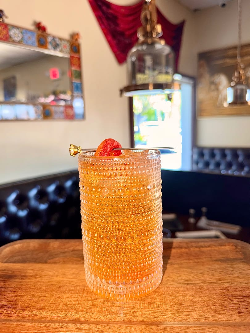 A strawberry-garnished cocktail in a textured glass sits on a wooden table in a restaurant.