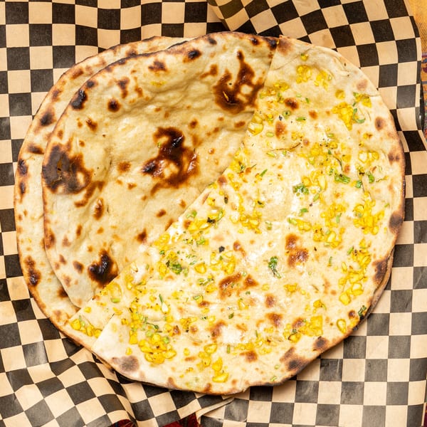 Two pieces of naan bread, one plain and one topped with corn and herbs, on checkered paper.