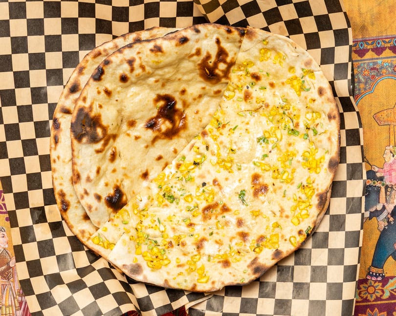 Two pieces of naan bread, one plain and one topped with corn and herbs, on checkered paper.