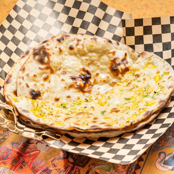 Two pieces of naan bread, one topped with corn and herbs, in a checkered paper-lined basket.