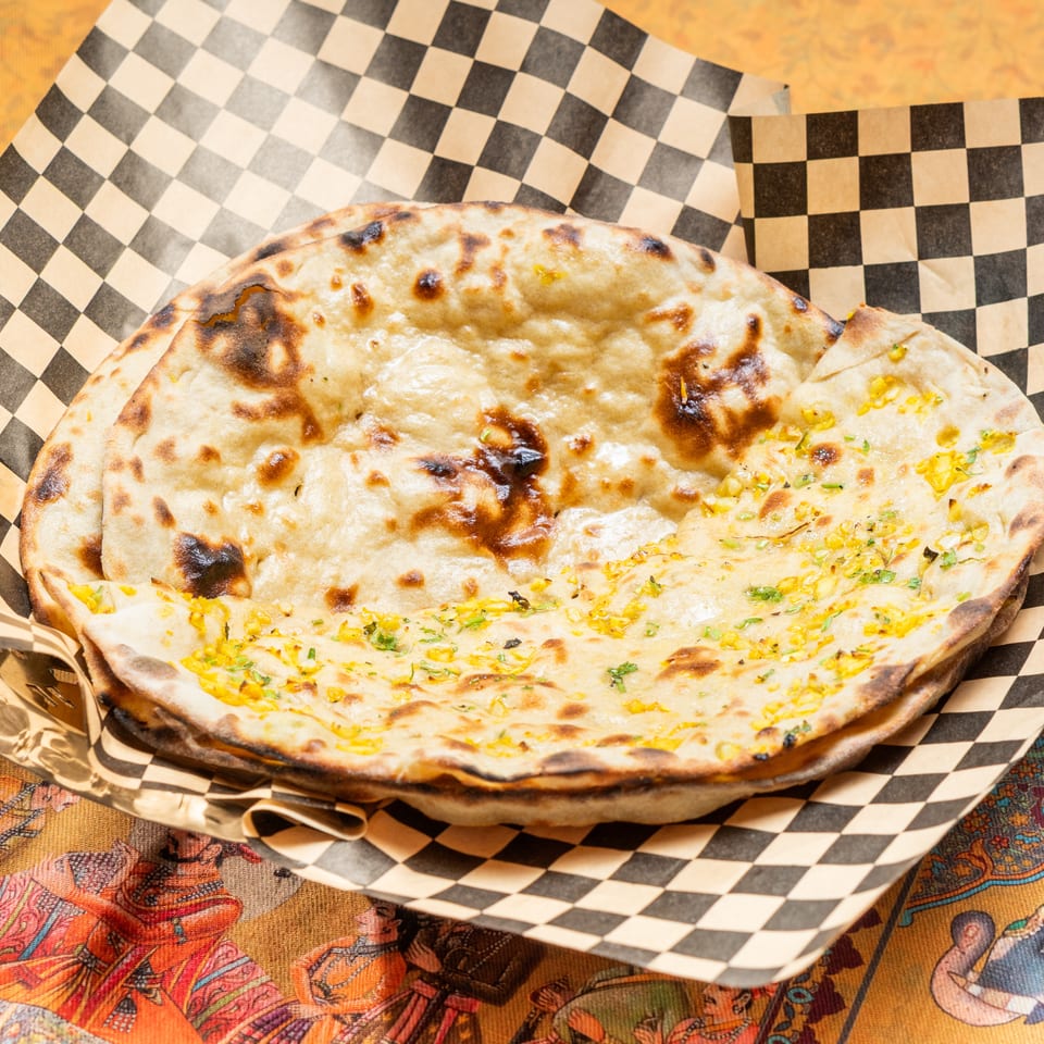 Two pieces of naan bread, one topped with corn and herbs, in a checkered paper-lined basket.