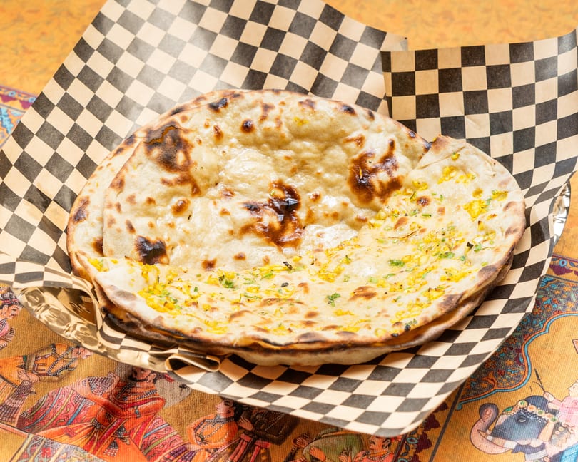 Two pieces of naan bread, one topped with corn and herbs, in a checkered paper-lined basket.