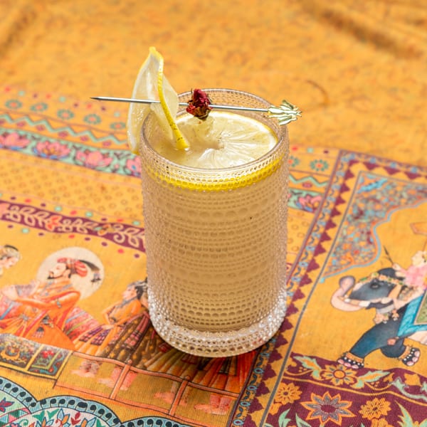 A refreshing lemon drink with a lemon slice and a dried rosebud garnish on a patterned yellow tablecloth.