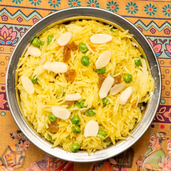 Yellow rice pilaf with green peas, golden raisins, and slivered almonds in a metal bowl.