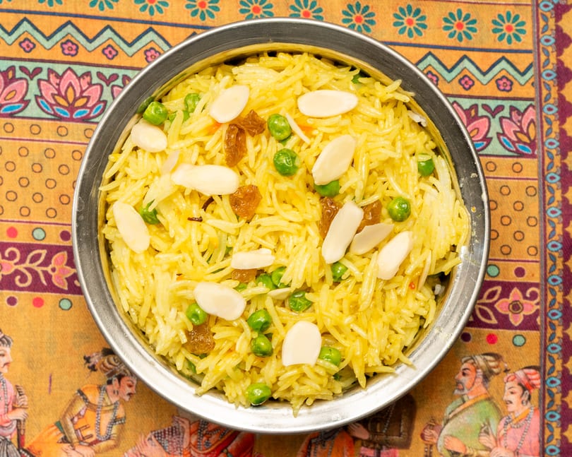 Yellow rice pilaf with green peas, golden raisins, and slivered almonds in a metal bowl.