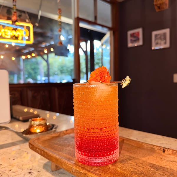 A vibrant orange and red cocktail with a flower garnish sits on a wooden tray in a restaurant.