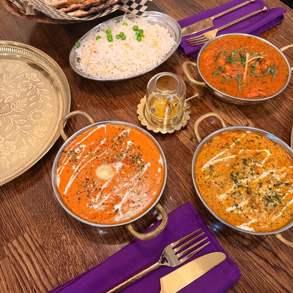 Three bowls of Indian curry, rice, and naan bread on a wooden table with purple napkins.