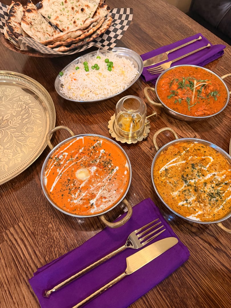 Three bowls of Indian curry, rice, and naan bread on a wooden table with purple napkins.