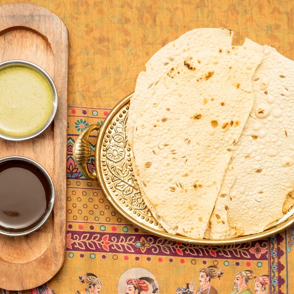 Crispy papadum with cumin seeds on a gold tray, served with two dipping sauces on a wooden board.