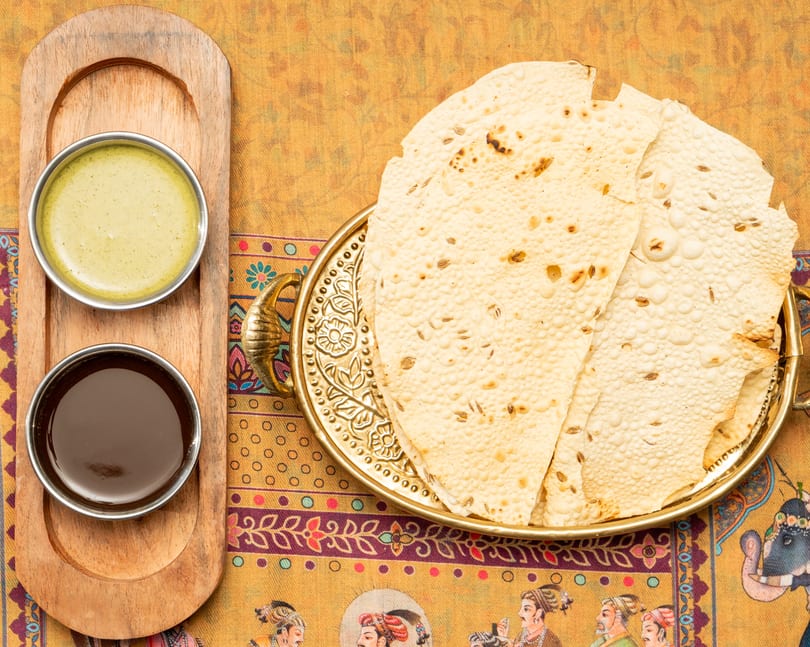 Crispy papadum with cumin seeds on a gold tray, served with two dipping sauces on a wooden board.