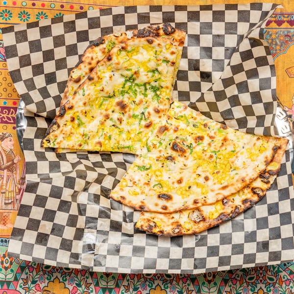 Two pieces of golden-brown naan bread with corn and herbs on checkered paper, on a colorful patterned tablecloth.