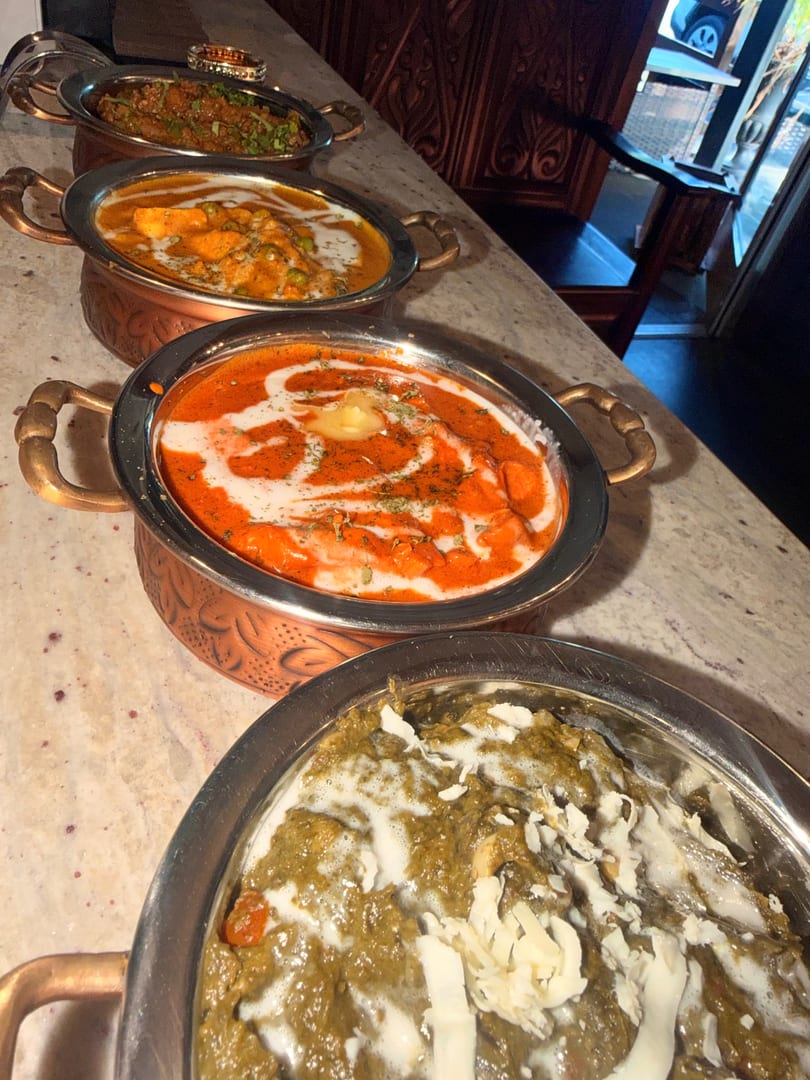 Four copper and silver bowls filled with various Indian curries, including green, orange, and red dishes.