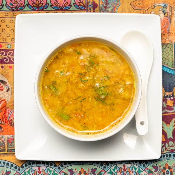 A bowl of yellow lentil soup with green herbs on a white plate with a spoon, on a colorful patterned tablecloth.