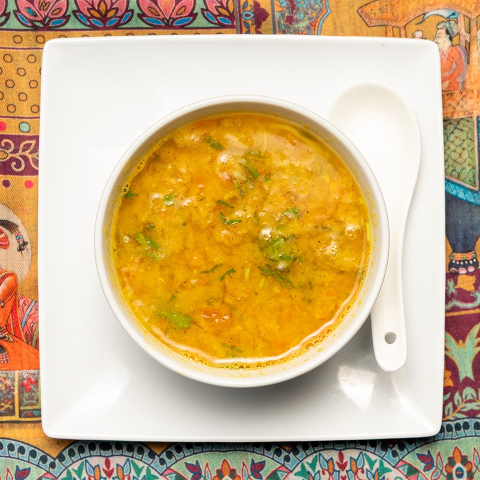 A bowl of yellow lentil soup with green herbs on a white plate with a spoon, on a colorful patterned tablecloth.