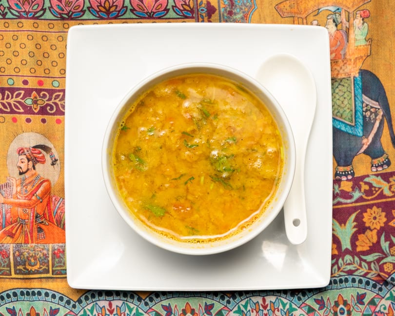 A bowl of yellow lentil soup with green herbs on a white plate with a spoon, on a colorful patterned tablecloth.