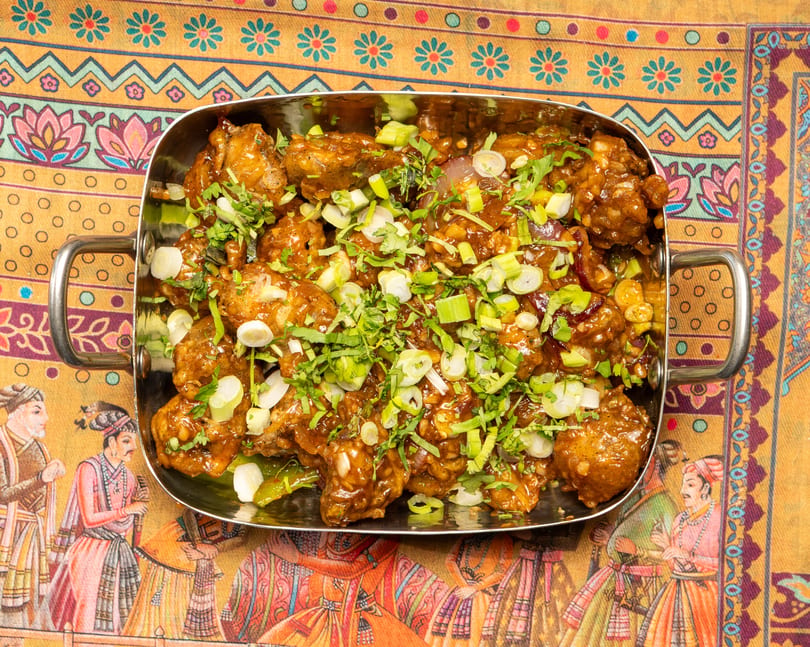 Chili chicken in a metal serving dish, garnished with green onions and cilantro, on a colorful patterned tablecloth.