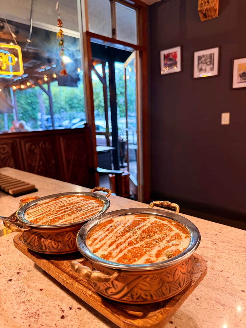 Two copper bowls of Indian curry with cream swirls on a wooden tray, with a restaurant interior and outdoor view.