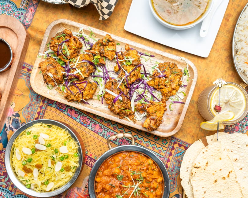 Overhead view of Indian dishes including pakoras, rice, chana masala, and papadum on a colorful tablecloth.