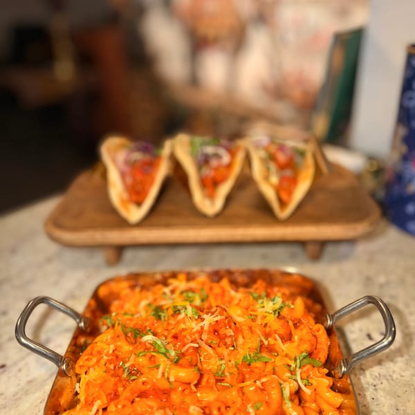 A metal pan of orange macaroni and cheese with herbs, and three tacos on a wooden board.