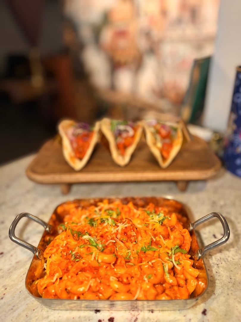 A metal pan of orange macaroni and cheese with herbs, and three tacos on a wooden board.