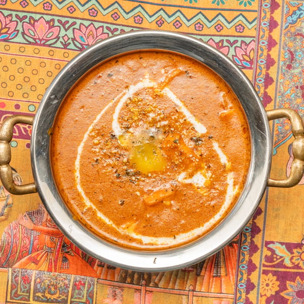 A metal bowl of orange curry with a swirl of cream and melted butter, on a colorful patterned tablecloth.