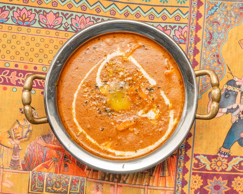 A metal bowl of orange curry with a swirl of cream and melted butter, on a colorful patterned tablecloth.
