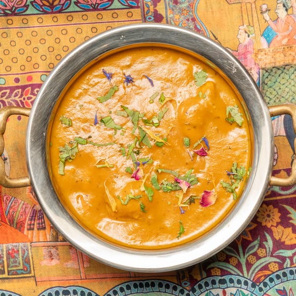 Indian curry in a metal bowl with brass handles, garnished with herbs and flower petals, on a colorful patterned tablecloth.
