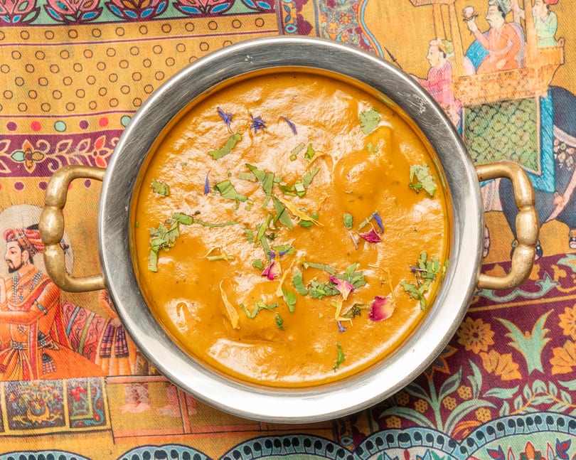 Indian curry in a metal bowl with brass handles, garnished with herbs and flower petals, on a colorful patterned tablecloth.